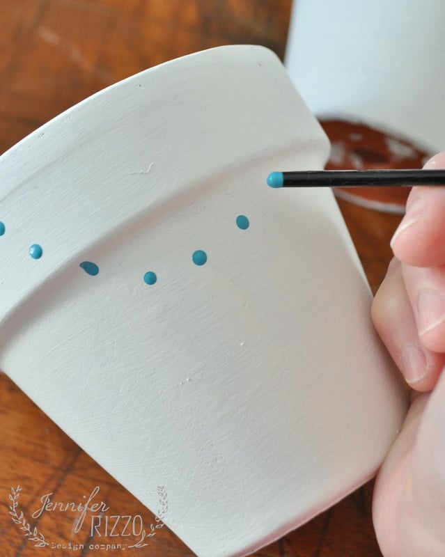 A hand uses a small brush to create paint dot flower pots, carefully applying blue dots onto a white terracotta pot.