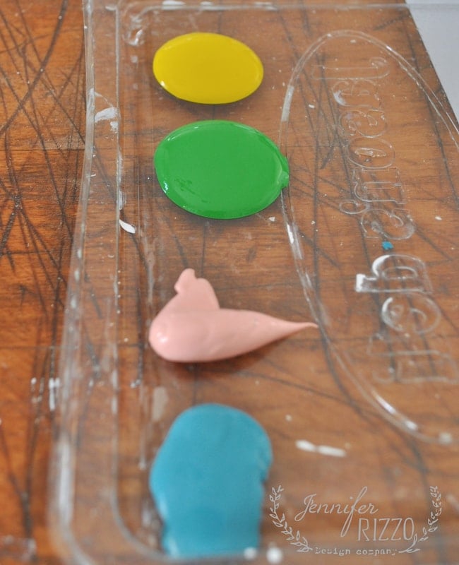A clear plastic tray on a wooden surface holds pools of yellow, green, pink, and blue paint—perfect for creating cheerful paint dot flower pots.