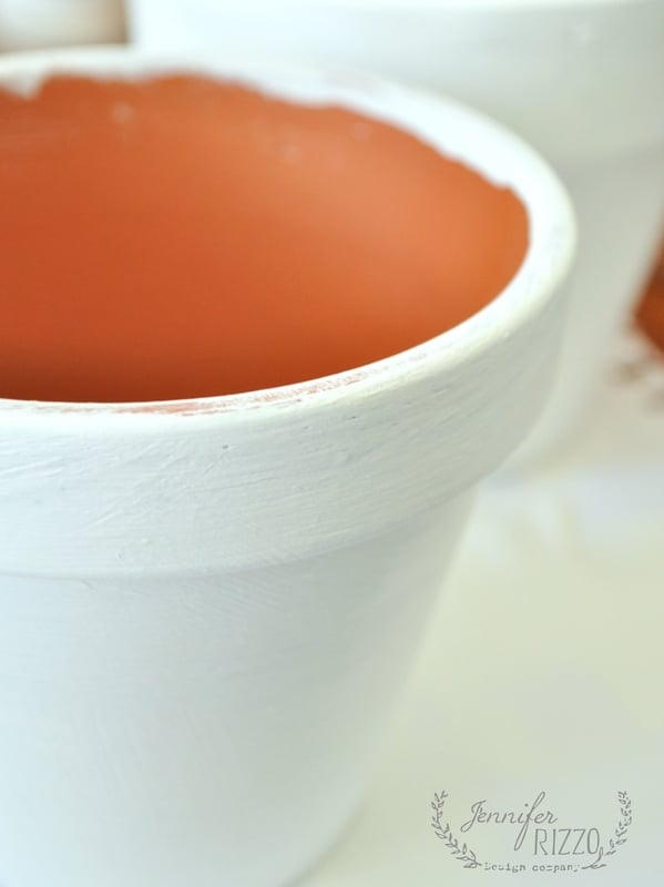 A close-up of a terracotta flower pot painted white on the outside, with the orange interior unpainted. This paint dot flower pot features "Jennifer Rizzo" faintly visible in the corner.