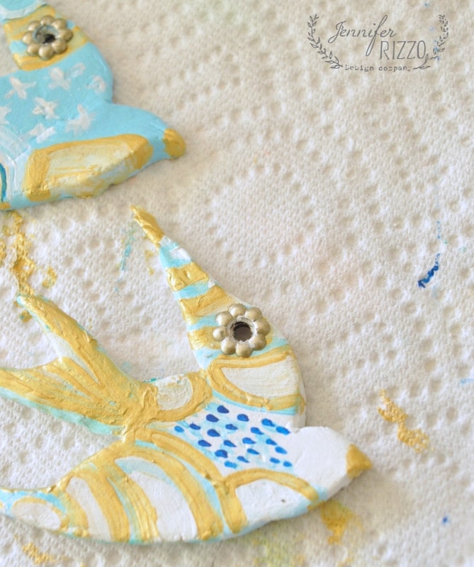 Two painted clay bird ornaments rest on a textured surface, adorned with blue, white, and gold patterns. Their intricate details echo the spirit of a DIY clay bird and feather wall hanging, bringing an artistic touch to any space.