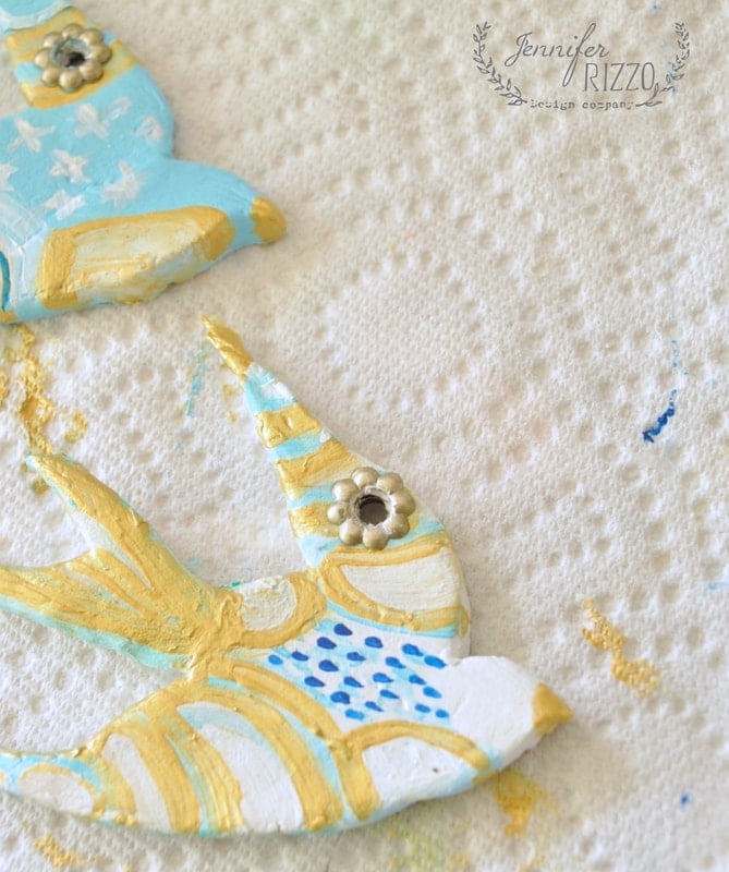 Two painted clay bird ornaments rest on a textured surface, adorned with blue, white, and gold patterns. Their intricate details echo the spirit of a DIY clay bird and feather wall hanging, bringing an artistic touch to any space.