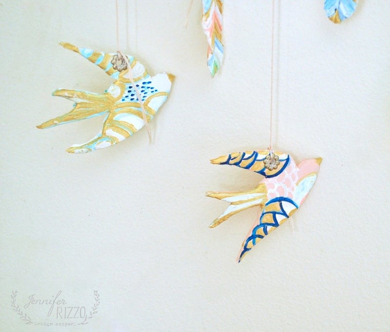 Three decorative, hand-painted clay bird ornaments hang from strings against a plain, light-colored wall, creating a charming DIY clay bird and feather wall hanging.