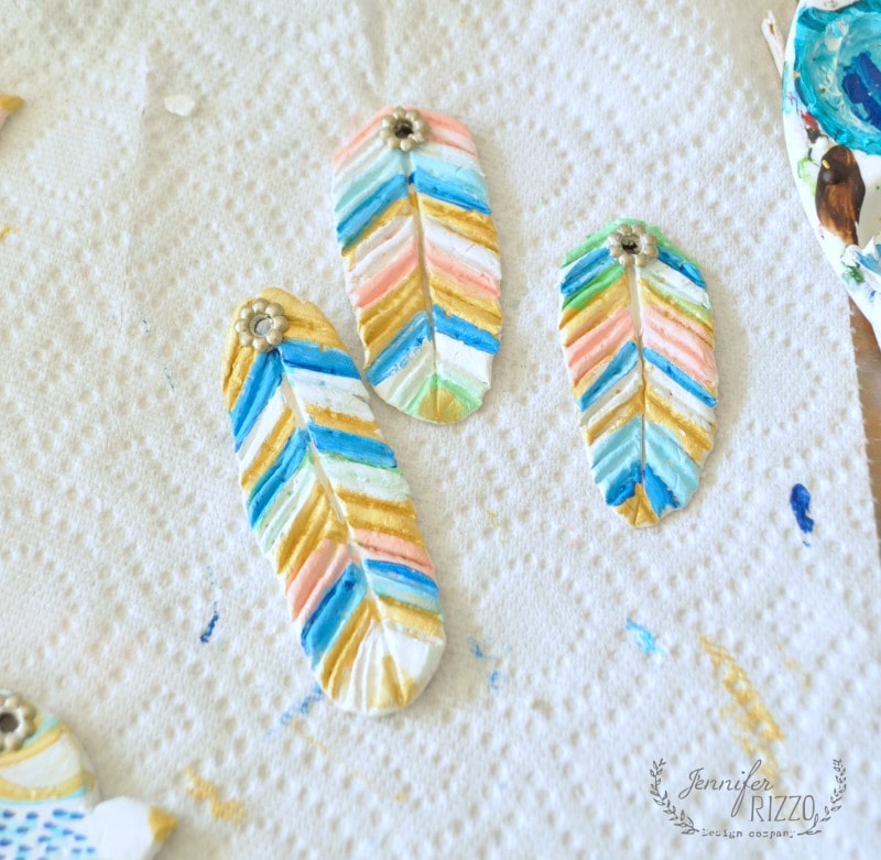 Painted feather-shaped clay ornaments for a DIY clay bird and feather wall hanging, adorned with colorful chevron patterns and metal flower embellishments, drying on a textured paper towel.