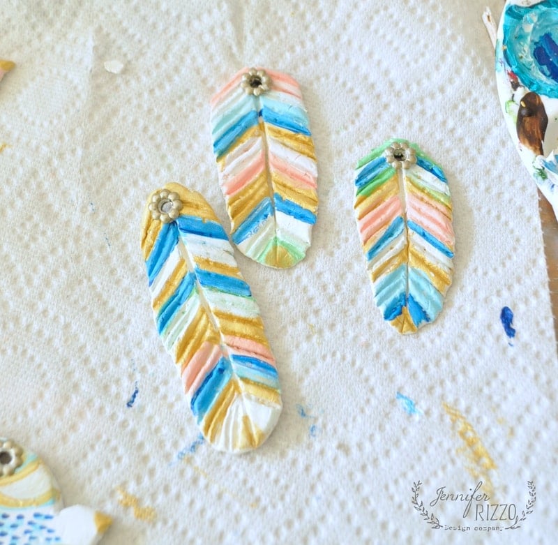 Painted feather-shaped clay ornaments for a DIY clay bird and feather wall hanging, adorned with colorful chevron patterns and metal flower embellishments, drying on a textured paper towel.