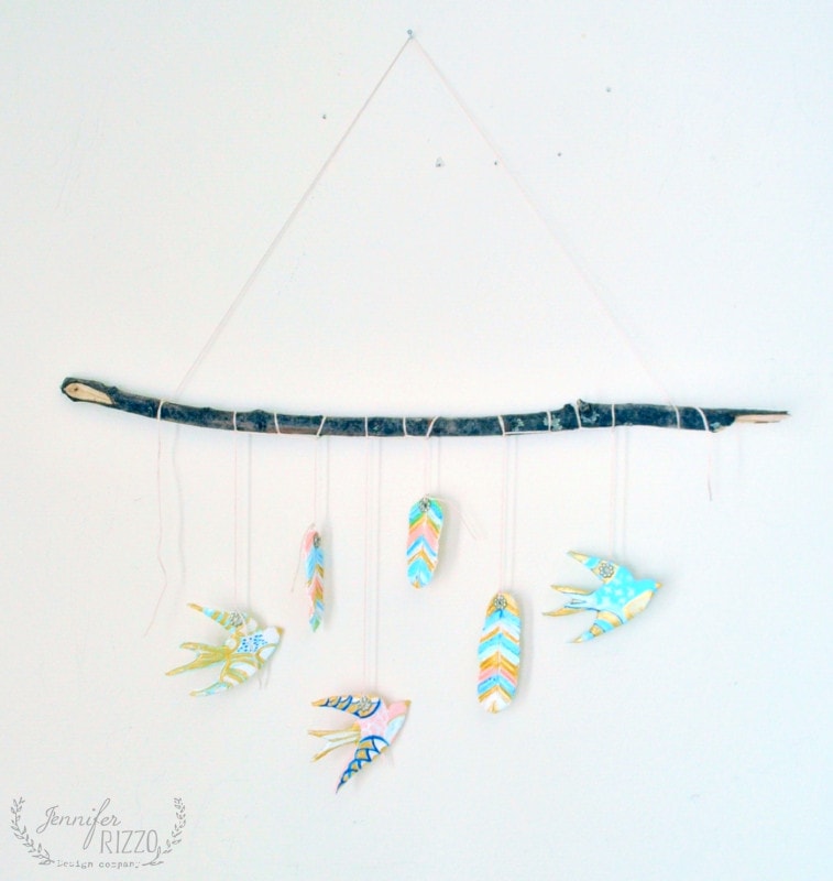 A handmade mobile features a DIY clay bird and feather wall hanging, with painted birds and feathers suspended from a horizontal stick against a white wall.