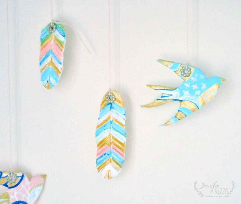 Hand-painted DIY clay bird and feather wall hanging, featuring ornaments in blue, pink, gold, and white, displayed against a plain white background.