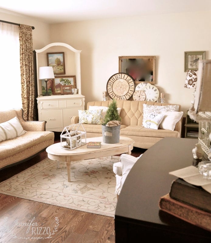 A cozy living room in this small house tour features beige furniture, a patterned rug, decorative clocks, framed art, and a potted plant on a white coffee table. Natural light streams through the curtained window.