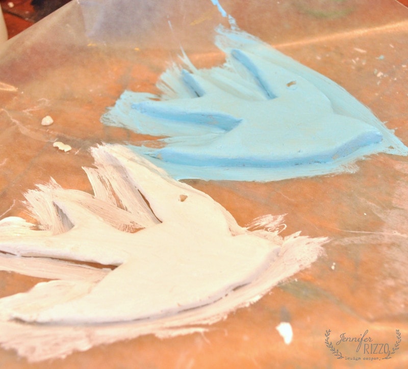 Two clay bird shapes, one painted blue and the other white, drying on a sheet of wax paper for a DIY clay bird and feather wall hanging.