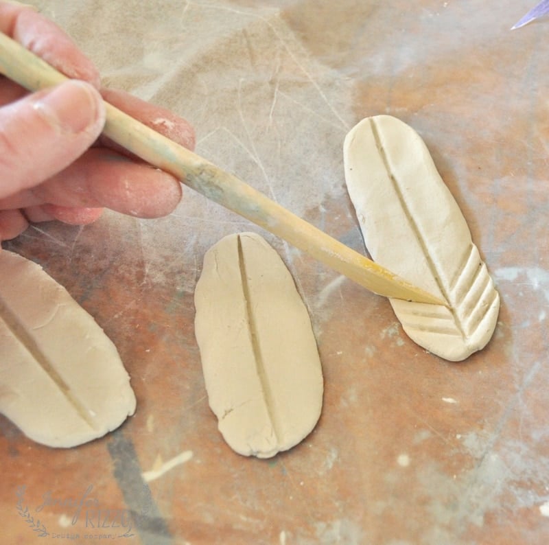A hand uses a wooden tool to carve feather-like lines into an oval piece of clay for a DIY clay bird and feather wall hanging, with two similar clay shapes nearby on a brown surface.