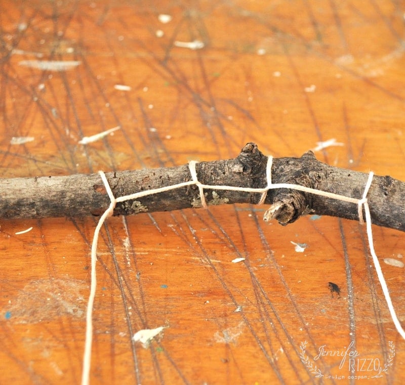A close-up of a stick tied with twine, resting on a scratched wooden surface—perfect for crafting a DIY clay bird and feather wall hanging.