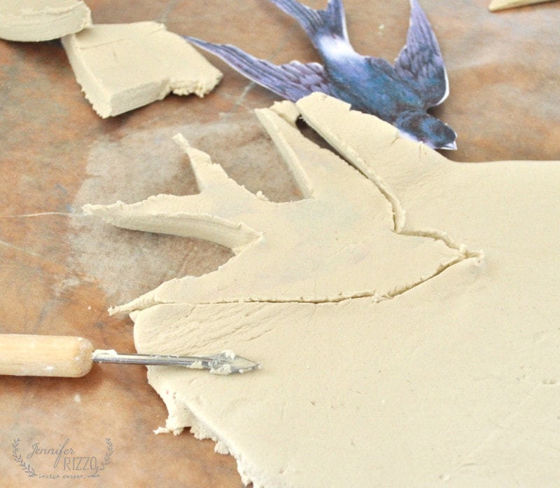 Air dry clay is being cut into the shape of a bird using a stencil and a sculpting tool on a work surface, perfect for creating your own DIY clay bird and feather wall hanging.