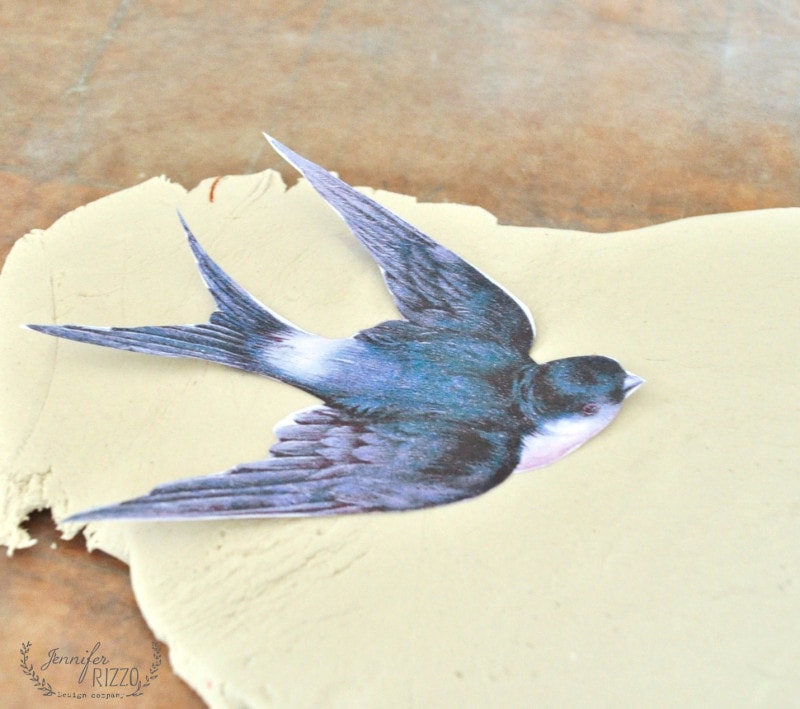 A paper cutout of a swallow is placed on rolled-out clay, the first step in creating a DIY clay bird and feather wall hanging on a wooden surface.