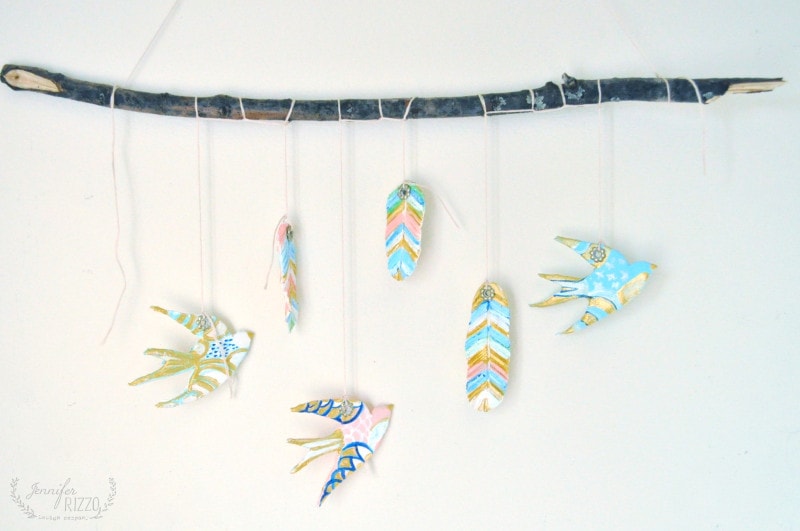 A handmade DIY clay bird and feather wall hanging features painted clay birds and feathers suspended from a horizontal branch by strings against a white background.