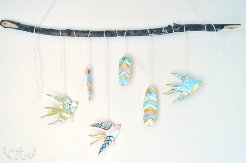 A handmade DIY clay bird and feather wall hanging features painted clay birds and feathers suspended from a horizontal branch by strings against a white background.