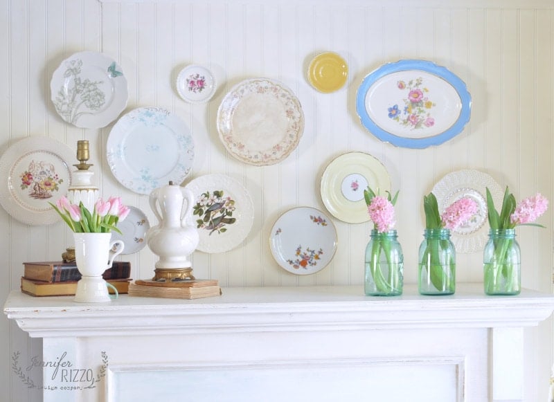 A white mantel decorated with stacked books, a white lamp, pink flowers in vases, and assorted decorative plates arranged on a white wall creates a charming Mother's Day party idea.