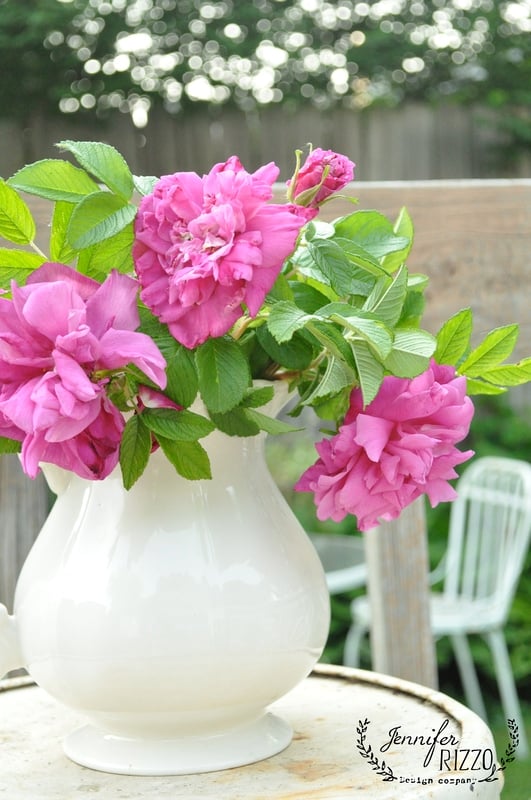 A white ceramic pitcher, serving as a charming example of creative plant and flower container ideas, is filled with bright pink roses and green leaves on an outdoor table, set against a blurred background of chairs and a fence.