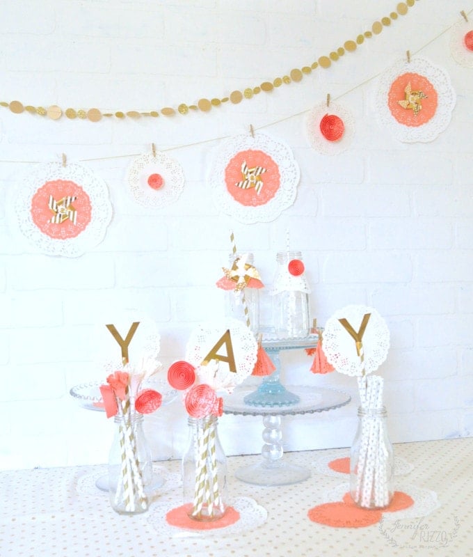 Table decorated with coral and gold themed party decorations, including doilies, paper straws, a Doily Paper Party Banner, vases with "YAY" letters, and a gold garland against a white brick wall.