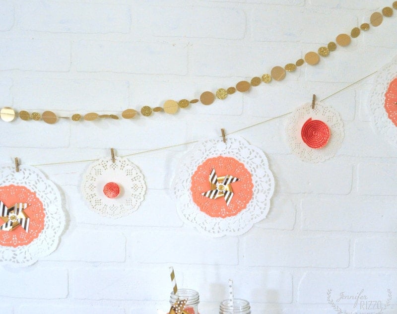 Two decorative garlands hang against a white brick wall; one is a Doily Paper Party Banner with paper doilies, coral circles, and small bows attached with clothespins, while the other features shiny gold circles.