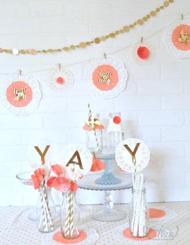 A pastel-themed party table is decorated with doilies, gold letter signs, glass bottles with straws, and a Doily Paper Party Banner garland against a white brick wall.
