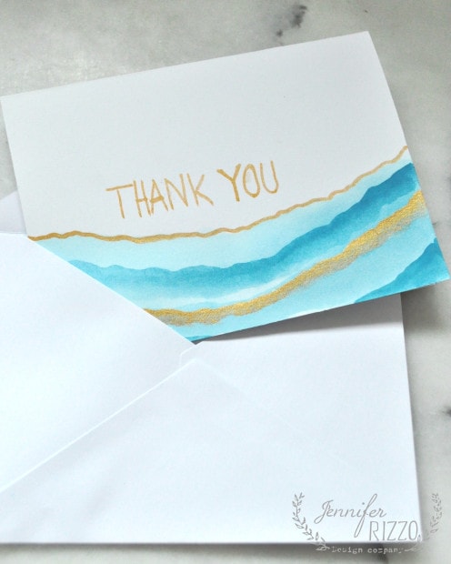 A thank you card with gold and blue watercolor stripes, partially pulled out of a white envelope, set on a light surface—perfect inspiration if you want to paint your own watercolor note cards.