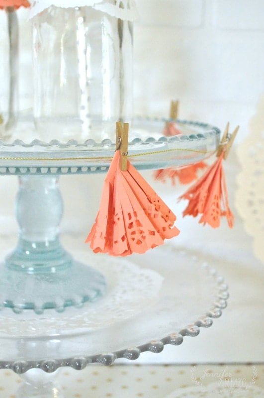 A glass cake stand decorated with coral paper tassels clipped on by small wooden clothespins, with paper doilies in the background, makes a charming scene—perfect alongside a doily paper party banner for your celebration.