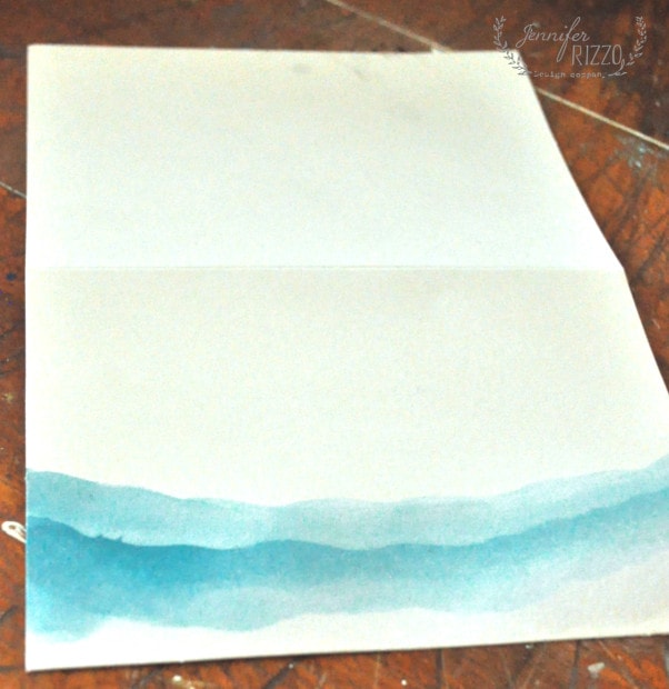 A piece of white paper is folded in half with a blue watercolor design along the bottom edge, set on a wooden surface—a simple invitation to paint your own watercolor note cards.