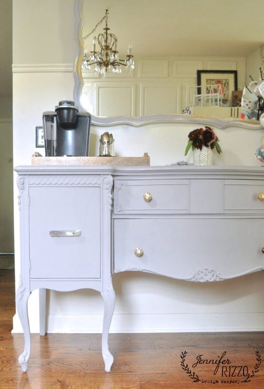 A vintage painted buffet in light gray with ornate details holds a Keurig coffee machine and a tray. There is a large mirror above, and a chandelier is partially visible in the reflection.