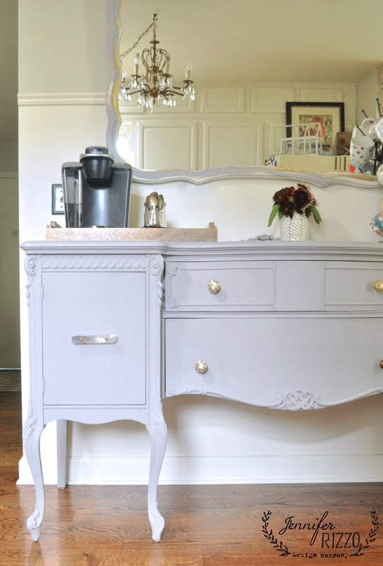 A vintage painted buffet in light gray with ornate details holds a Keurig coffee machine and a tray. There is a large mirror above, and a chandelier is partially visible in the reflection.