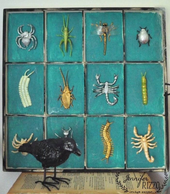 A display box with twelve toy insects and arachnids, arranged in a 3x4 grid, evokes the atmosphere of a creepy mad scientist Halloween laboratory. A black bird figurine stands in front on a closed book, all set against a teal background.