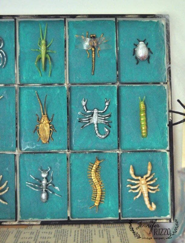 A display case with nine compartments, each containing a different metallic model of an insect or arachnid on a teal background—perfect for adding a touch of creepy mad scientist Halloween laboratory flair to your décor.