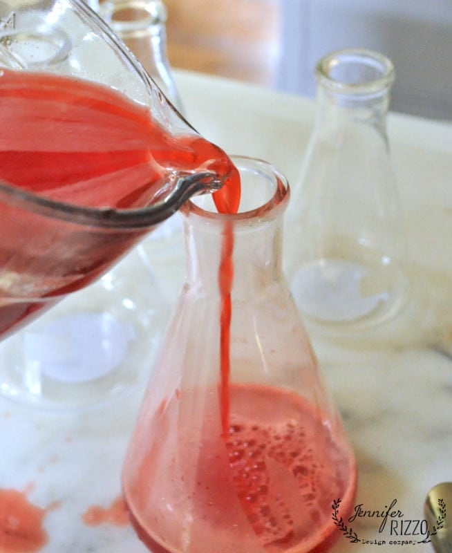 Red liquid is being poured from a glass pitcher into a laboratory flask on a white surface, evoking the feel of a creepy mad scientist Halloween laboratory, with empty flasks looming in the background.