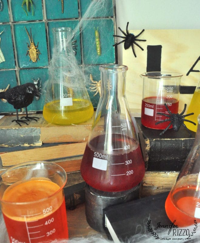 Various colored liquids in beakers and flasks are arranged among Halloween decorations, including fake spiders, a black bird, and cobwebs, creating the perfect setting for a creepy mad scientist Halloween laboratory on a table with books.