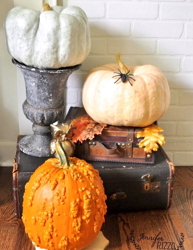 Three pumpkins of different colors and textures are arranged on vintage suitcases, accented with a fake spider and autumn leaves—perfect Halloween mantel decor for a cozy seasonal display.
