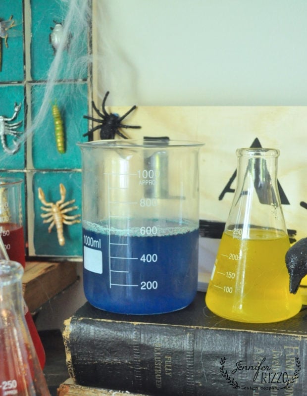 A beaker with blue liquid and a flask with yellow liquid sit on old books, surrounded by plastic insects and Halloween decorations, creating the perfect creepy mad scientist Halloween laboratory scene.