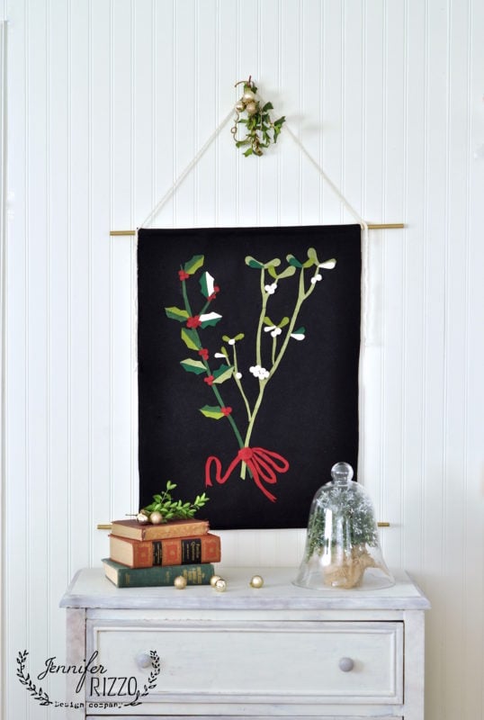 A fabric wall hanging with a mistletoe design is displayed above a dresser with stacked books, including the holiday book Creatively Christmas, a plant under a glass cloche, and gold ornaments.
