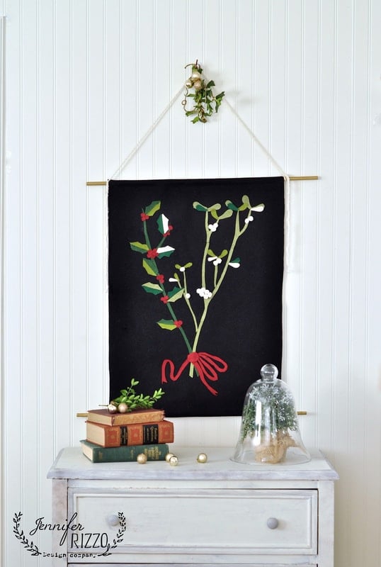 A fabric wall hanging with a mistletoe design is displayed above a dresser with stacked books, including the holiday book Creatively Christmas, a plant under a glass cloche, and gold ornaments.