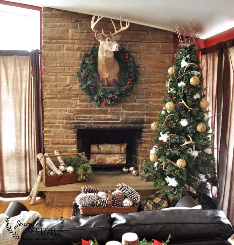 A living room with a stone fireplace, deer head mount with wreath, decorated Christmas tree, and baskets of pinecones and logs—like a scene from the holiday book Creatively Christmas.