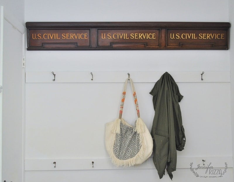 A wall-mounted rack on white painted walls features three sections labeled "U.S. Civil Service," with hooks below holding a patterned tote bag and a green jacket.