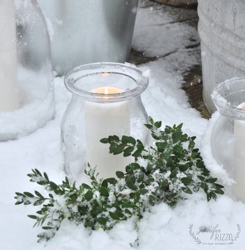 A white candle in a glass holder sits on snow beside green leafy branches, creating a cozy holiday outdoor scene with snow-covered metal containers in the background.