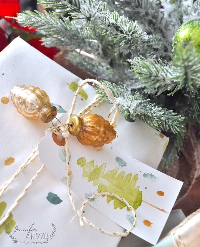 A wrapped gift with white and green DIY gift wrap, decorated with a gold ornament, a string, and a tag featuring a watercolor Christmas tree next to a frosted evergreen branch.