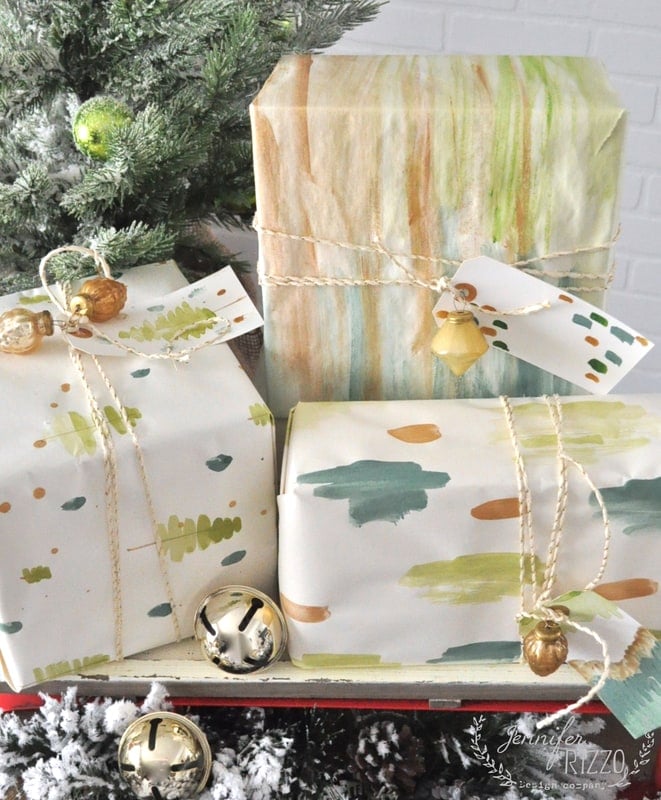 Three presents wrapped in DIY gift wrap featuring abstract, watercolor-patterned paper with twine and small gold ornaments, displayed near a Christmas tree.