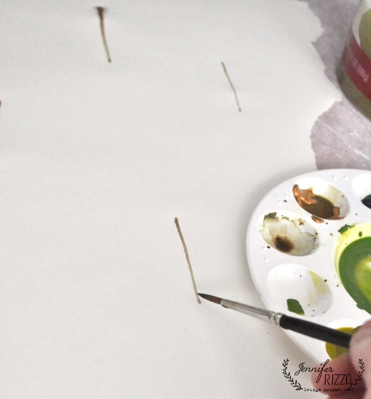 A hand uses a paintbrush to make vertical brown lines on white paper next to a paint palette with green and brown paint, creating unique DIY gift wrap.