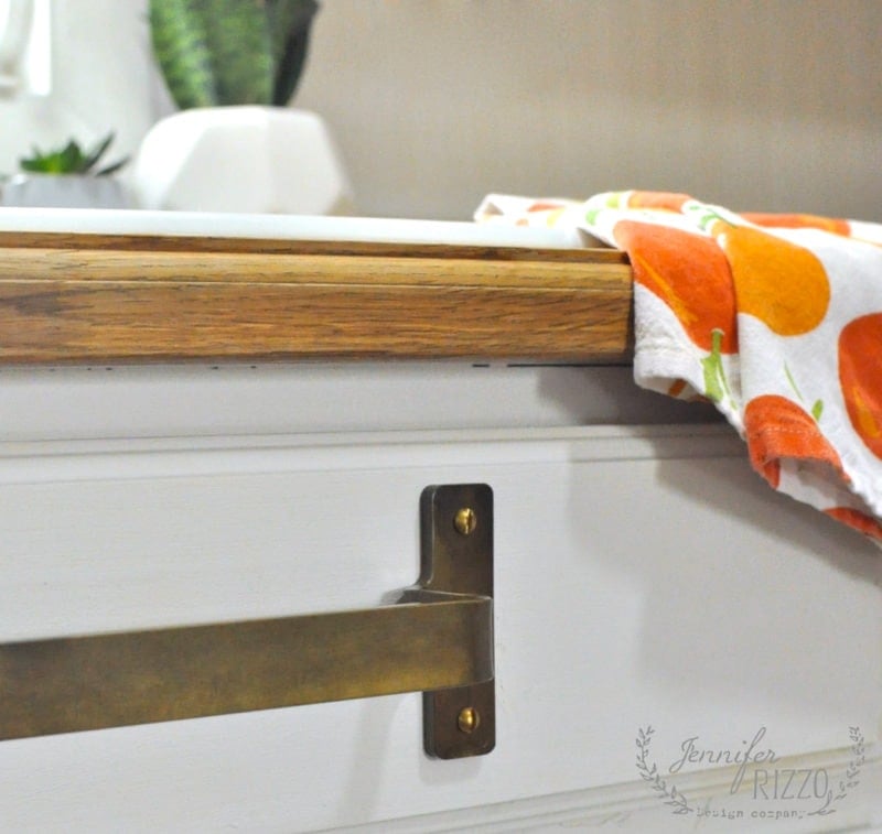 Close-up of a kitchen drawer with a brass towel bar handle, topped with a wood countertop and an orange-patterned towel, with a potted plant blurred in the background.