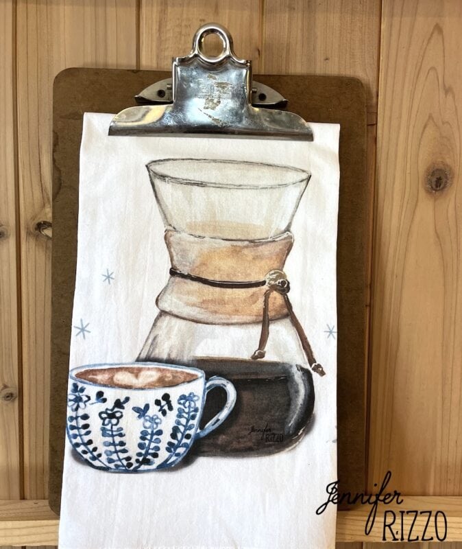 A tea towel on a clipboard features an illustration of a Chemex coffee maker and a blue patterned coffee cup, set against a wooden background—perfect for display on a brass towel bar.