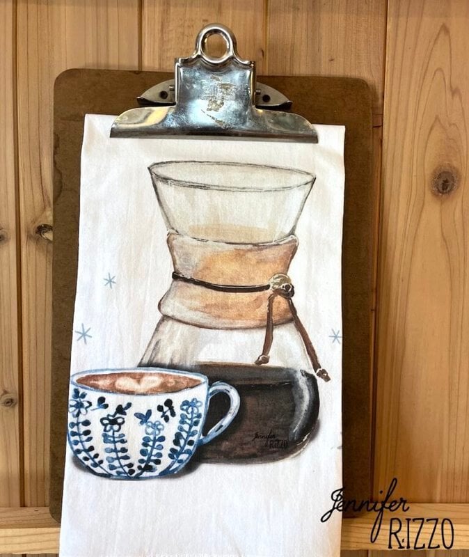 A tea towel on a clipboard features an illustration of a Chemex coffee maker and a blue patterned coffee cup, set against a wooden background—perfect for display on a brass towel bar.