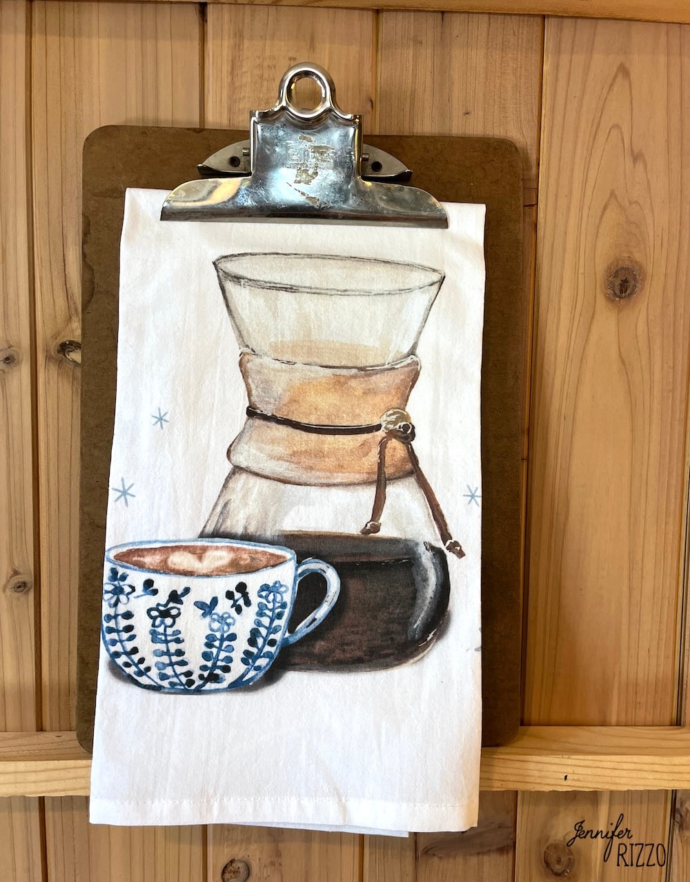 A tea towel on a clipboard features an illustration of a Chemex coffee maker and a blue patterned coffee cup, set against a wooden background—perfect for display on a brass towel bar.