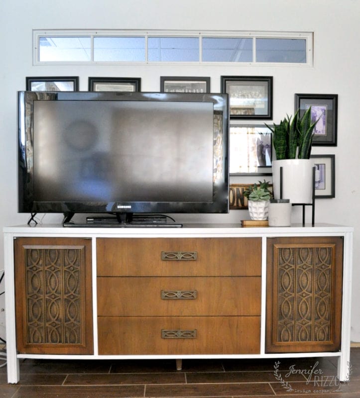 A flat-screen TV sits on a Mid Century Modern furniture makeover console with three central drawers and two patterned side cabinets; framed photos and potted plants are arranged behind and beside the TV.