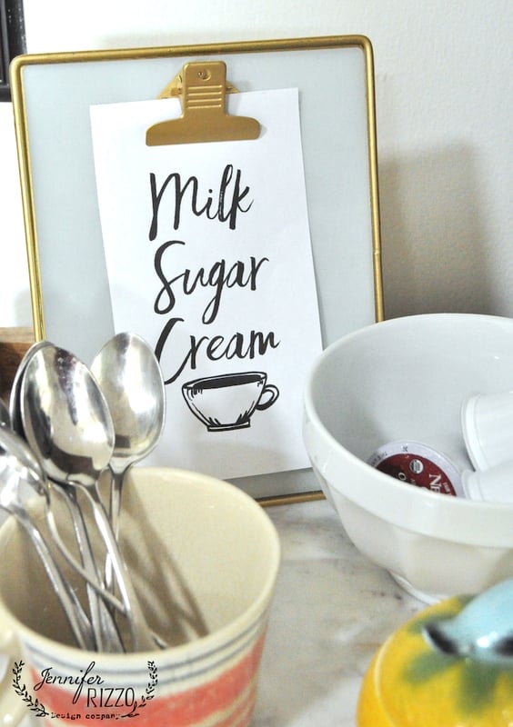 A countertop display with a sign reading "Milk Sugar Cream" sits in front of classic kitchen subway tile, next to bowls containing spoons and coffee pods.