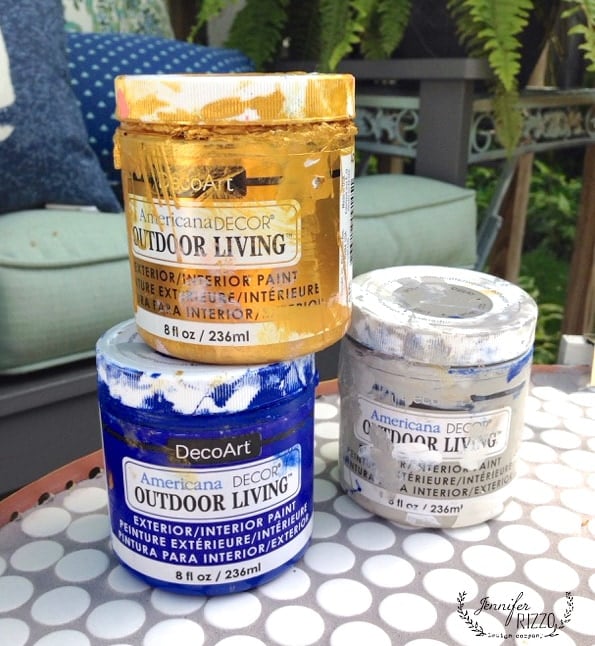 Three jars of DecoArt Americana Decor Outdoor Living paint in gold, white, and blue sit on a round, patterned outdoor table—perfect for your next DIY penny tile table top project—with patio furniture in the background.