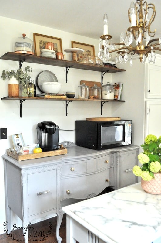 Gray sideboard with a black microwave, coffee maker, and assorted jars beneath two wooden shelves displaying dishes, plants, and framed art. Kitchen subway tile adds a fresh touch to this bright kitchen with a striking chandelier.